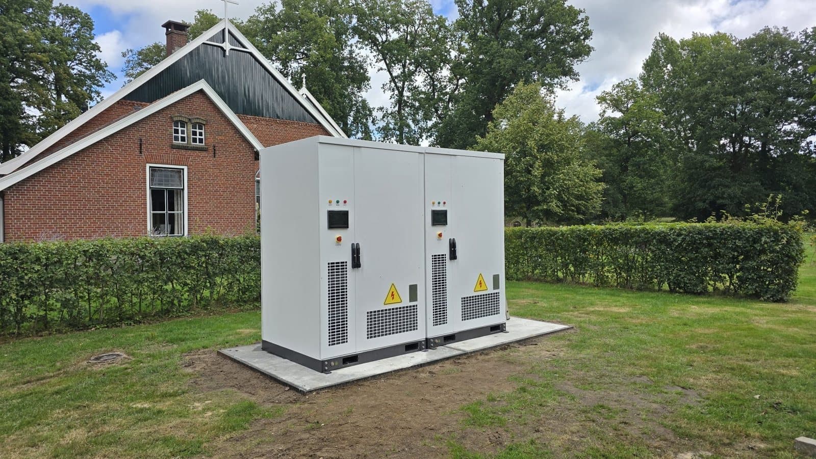 Twee batterijkasten op een betonplaat in het gras naast een boerderij